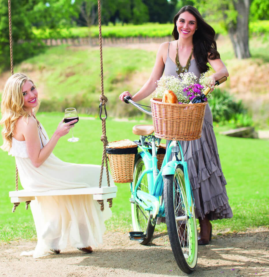 Woman on swing holding a glass of wine and woman standing next to a bike.